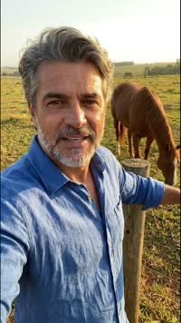 Fazendeiro de camisa de linho azul falando na cerca, com o cavalo atrás, no fim da tarde